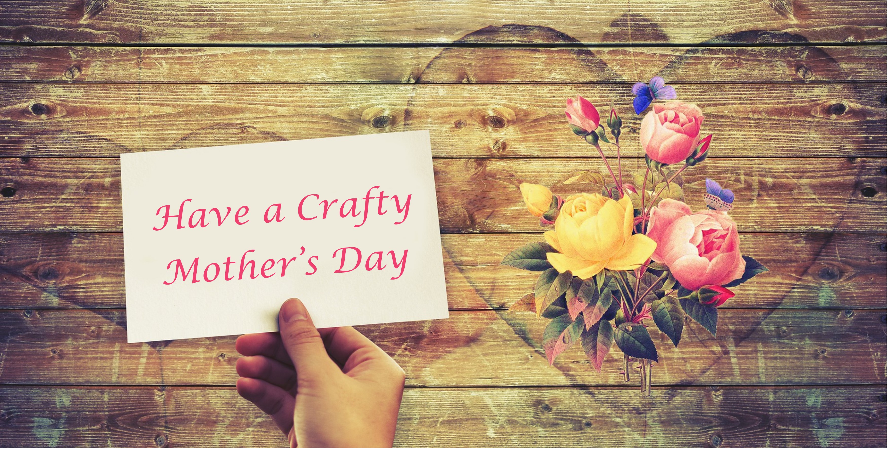 Hand holding a card with the wording Have a Crafty Mother's Day in pink writing, above a wooden background with a rose bouquet in a heart shape to the right